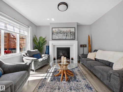 Living area featuring a contemporary fireplace with a white mantel, light wood-finish flooring, a large window, a ceiling-mounted light fixture, and light-toned walls - 323 Balsam Link, Leduc, AB - Indoor Photo Showing Living Room With Fireplace