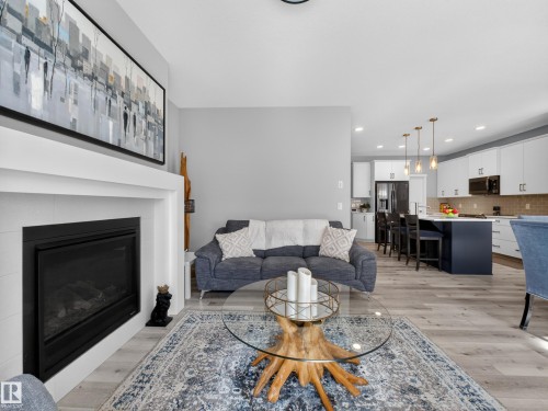 Open-concept living area featuring a modern fireplace, wood-finish flooring, recessed lighting, and a kitchen with white cabinetry, stainless steel appliances, and a dark blue island with pendant lighting - 323 Balsam Link, Leduc, AB - Indoor Photo Showing Living Room With Fireplace