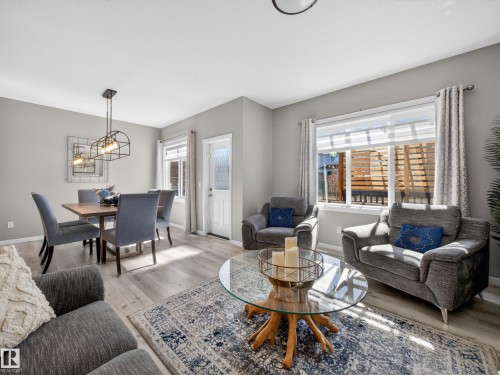 Open-concept living area featuring light wood-finish flooring, a modern linear chandelier above the dining space, ample natural light from multiple windows, a white paneled entry door with decorative glass, and light gray wall tones - 323 Balsam Link, Leduc, AB - Indoor Photo Showing Living Room