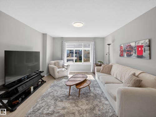 Light-filled living area featuring wood-finish flooring, a contemporary ceiling light fixture, a large window with blinds, and light-toned wall finishes - 323 Balsam Link, Leduc, AB - Indoor Photo Showing Living Room