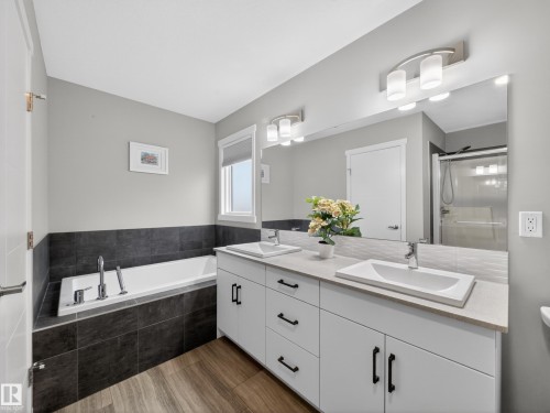 Bathroom featuring a white double vanity with rectangular undermount sinks, a built-in bathtub with dark tile surround, a large frameless mirror, and wood-finish flooring - 323 Balsam Link, Leduc, AB - Indoor Photo Showing Bathroom