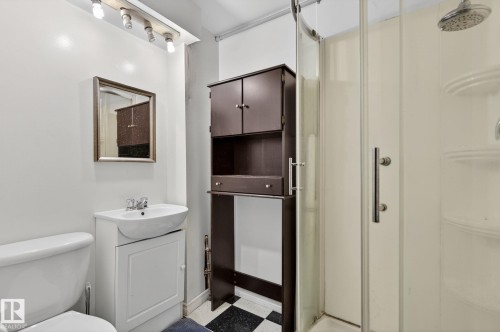 Compact bathroom featuring a glass-enclosed shower, white ceramic toilet, vanity with integrated basin, dark wood-finish storage tower, and checkerboard floor tiling - 9112 130 Avenue, Edmonton, AB - Indoor Photo Showing Bathroom