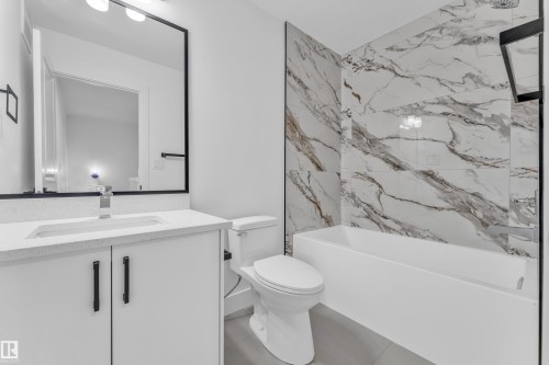 Contemporary bathroom featuring a vanity with a white countertop and integrated sink, a large mirror with a black frame, a white toilet, and a bathtub with marble-look wall tiling - 277 Munn Way, Leduc, AB - Indoor Photo Showing Bathroom