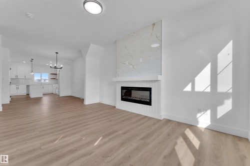 Spacious open-concept interior featuring light wood-finish flooring, a modern fireplace with a white shiplap base and a stone-look surround, and a kitchen area with white cabinetry and an island - 285 Munn Way, Leduc, AB - Indoor Photo Showing Living Room With Fireplace