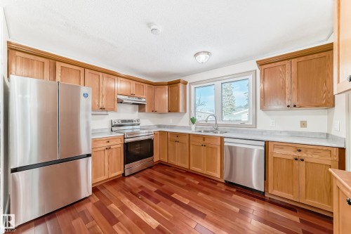 The kitchen features wood cabinetry, stainless steel appliances, light-colored countertops, and hardwood flooring - 8733 181A Street Nw, Edmonton, AB - Indoor Photo Showing Kitchen With Double Sink