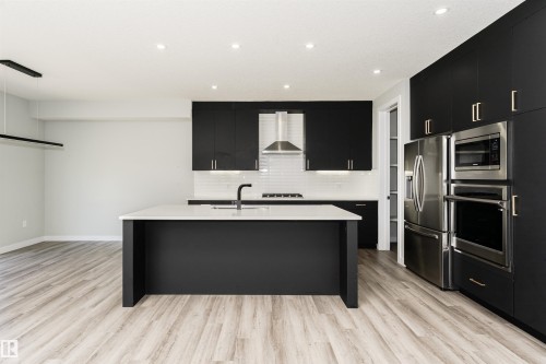 Contemporary kitchen featuring dark flat-panel cabinetry, light wood-finish flooring, a central island with a white countertop, stainless steel appliances, and a white subway tile backsplash - 5386 Kimball Place, Edmonton, AB - Indoor Photo Showing Kitchen