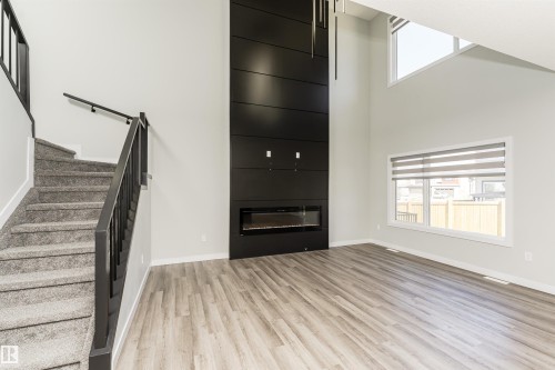 Two-story living area featuring a black paneled fireplace surround with an electric insert, high ceilings, large windows with blinds, wood-finish flooring, and a carpeted staircase with black railings - 5386 Kimball Place, Edmonton, AB - Indoor With Fireplace