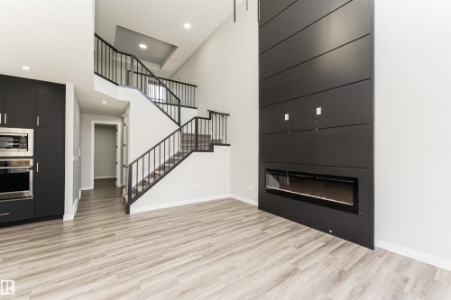 Vaulted ceilings enhance the open-concept design featuring a contemporary fireplace with horizontal paneling, a built-in oven and microwave, light wood-finish flooring, and a two-story staircase with black metal railings - 5386 Kimball Place, Edmonton, AB - Indoor With Fireplace