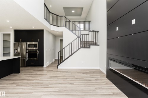 Open-concept living area featuring light wood-finish flooring, a modern black fireplace, and a two-story staircase with black metal railings - 5386 Kimball Place, Edmonton, AB - Indoor Photo Showing Other Room