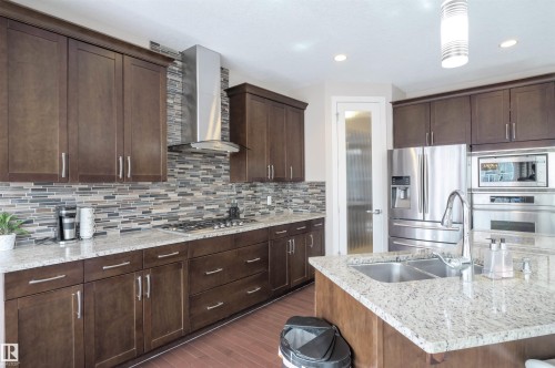 Contemporary kitchen featuring rich wood cabinetry, granite countertops, a mosaic tile backsplash, stainless steel appliances, and a central island with a double basin sink - 1728 53 Street, Edmonton, AB - Indoor Photo Showing Kitchen With Double Sink With Upgraded Kitchen