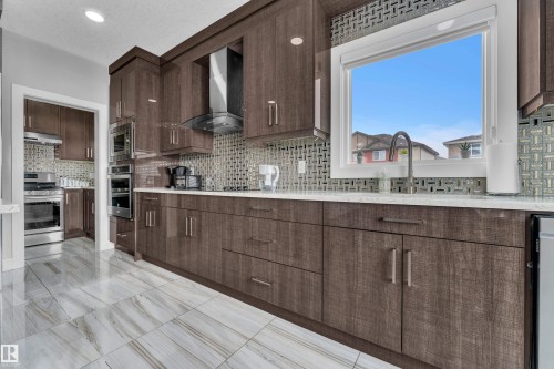 Kitchen featuring wood-finish cabinetry, stainless steel appliances, a unique tile backsplash, a stainless steel sink, and light-toned stone countertops - 1404 25 Street, Edmonton, AB - Indoor Photo Showing Kitchen