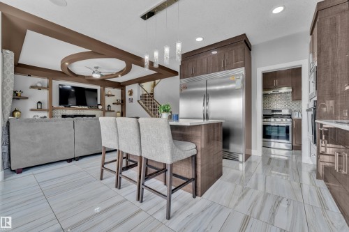 Open-concept living space featuring sleek tile flooring, a kitchen island with seating, dark wood-finish cabinetry, a built-in stainless steel refrigerator, and a recessed ceiling with decorative beams - 1404 25 Street, Edmonton, AB - Indoor