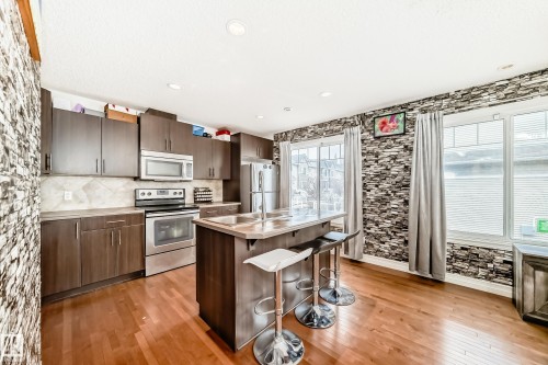 Kitchen featuring an island with breakfast bar seating, stainless steel appliances, wood-finish flooring, dark wood cabinetry, and a stone-look accent wall - 69 3625 144 Avenue, Edmonton, AB - Indoor Photo Showing Kitchen