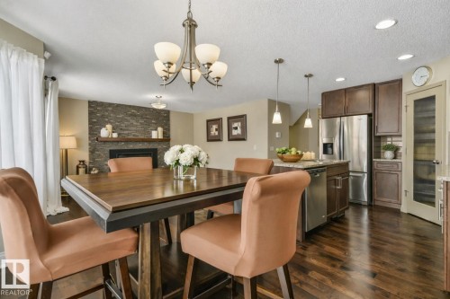 Open-concept living space featuring wood-finish flooring, a stacked stone fireplace, a kitchen island with pendant lighting, stainless steel appliances, and dark wood cabinetry - 21851 80 Avenue, Edmonton, AB - Indoor Photo Showing Dining Room