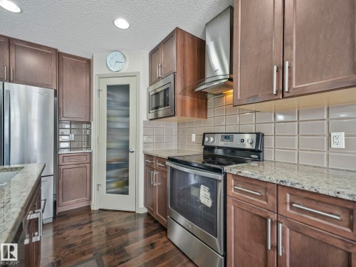 Kitchen featuring wood-finish cabinetry, stainless steel appliances, light granite countertops, a tiled backsplash, and dark wood-finish flooring - 21851 80 Avenue, Edmonton, AB - Indoor Photo Showing Kitchen With Upgraded Kitchen