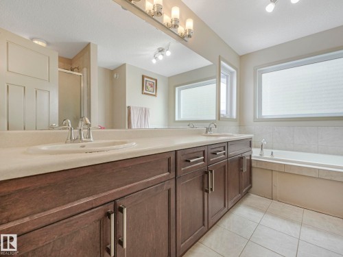 Double vanity with integrated sinks and brushed nickel faucets, dark wood-finish cabinetry, broad wall mirror, recessed bathtub with tiled surround, and white tiled flooring - 21851 80 Avenue, Edmonton, AB - Indoor Photo Showing Bathroom