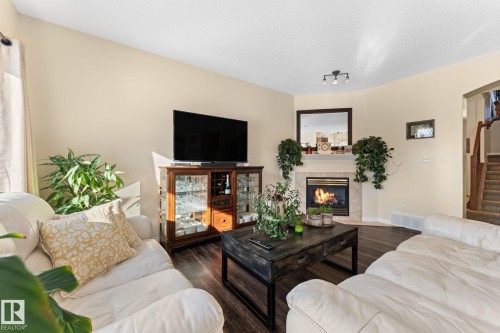Living area featuring dark flooring, a corner fireplace with a light-colored surround, and a staircase with wooden handrails - 112 Lakeland Point(E), Beaumont, AB - Indoor Photo Showing Living Room With Fireplace