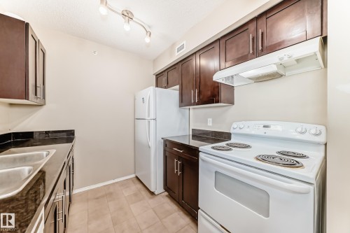 Kitchen featuring dark wood-finish cabinetry, dark countertops, a double basin stainless steel sink, and a white electric range with an overhead exhaust fan - 404 12025 22 Avenue Sw, Edmonton, AB - Indoor Photo Showing Kitchen