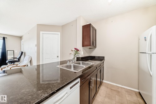 Kitchen area featuring a double basin stainless steel sink, dark cabinetry, and a white refrigerator - 404 12025 22 Avenue Sw, Edmonton, AB - Indoor Photo Showing Kitchen With Double Sink