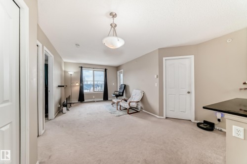 Spacious living area featuring neutral carpeting, a large window with dark drapes, and a dropped ceiling light fixture - 404 12025 22 Avenue Sw, Edmonton, AB - Indoor Photo Showing Other Room