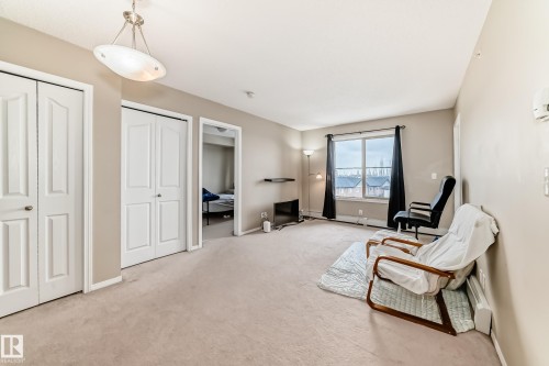 Carpeted living space featuring neutral wall paint, a large window, a ceiling-mounted light fixture, and two white bi-fold closet doors - 404 12025 22 Avenue Sw, Edmonton, AB - Indoor