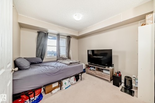 Carpeted interior room with neutral-toned walls, featuring a window with dark curtains and a ceiling-mounted light fixture - 404 12025 22 Avenue Sw, Edmonton, AB - Indoor Photo Showing Bedroom