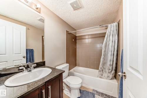 Bathroom featuring a dark wood vanity with a stone-look countertop and an integrated sink, a full-size mirror with vanity lighting, a toilet, and a bathtub with tiled surround - 404 12025 22 Avenue Sw, Edmonton, AB - Indoor Photo Showing Bathroom