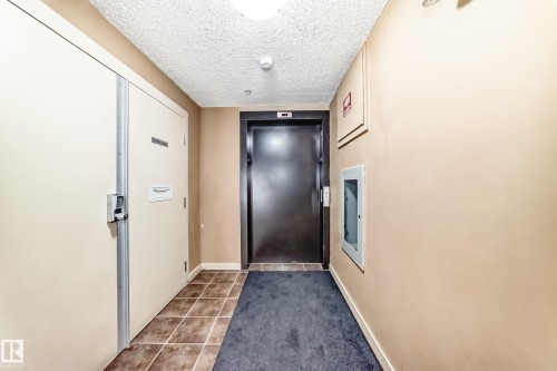 Corridor featuring a dark-paneled elevator, neutral wall paint, textured ceiling, and mixed flooring with large format tiles and carpeting - 404 12025 22 Avenue Sw, Edmonton, AB - Indoor Photo Showing Other Room