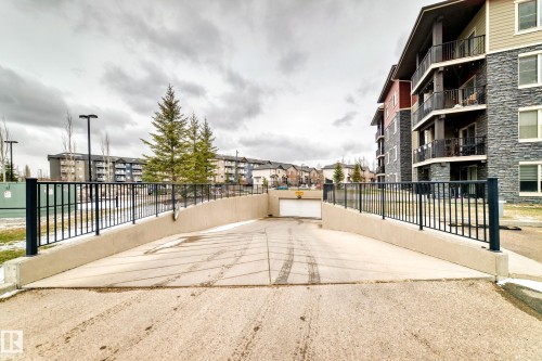 Concrete ramp leading to an underground parking garage, bordered by black metal railings and a stone-clad building exterior featuring multiple balconies - 404 12025 22 Avenue Sw, Edmonton, AB - Outdoor With Balcony