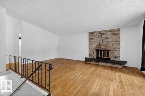 Spacious room featuring wood-finish flooring, a stone-faced fireplace with a dark hearth, white walls, and a textured ceiling - 6018 105A Street, Edmonton, AB 