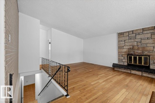 Spacious room featuring wood-finish flooring, a stone-clad fireplace with a black hearth, and a white brick accent wall - 6018 105A Street, Edmonton, AB 