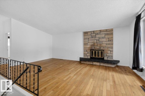 Spacious room featuring wood-finish flooring, a stone-clad fireplace with a black hearth, and a large window providing natural light - 6018 105A Street, Edmonton, AB 