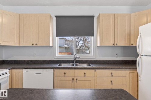 Kitchen featuring light wood-finish cabinetry, dark countertops, a double basin stainless steel sink, a white dishwasher, and a white refrigerator - 1904 36 Avenue, Edmonton, AB - Indoor Photo Showing Kitchen With Double Sink