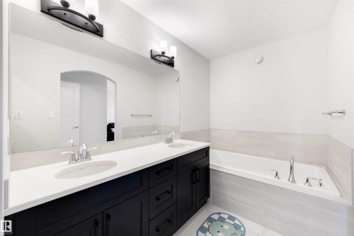 Spacious bathroom featuring a double vanity with white countertops and dark cabinetry, a large integrated mirror, and an undermount bathtub with tiled surround - 3193 Checknita Way, Edmonton, AB - Indoor Photo Showing Bathroom