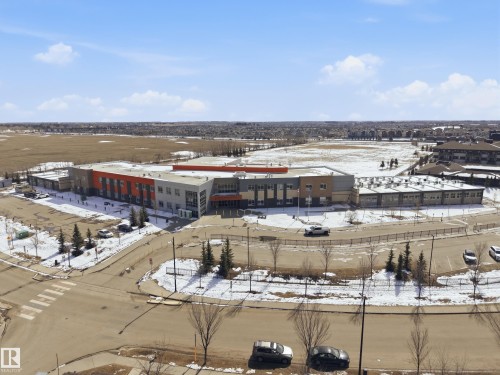 Aerial view of the community, featuring a modern building with a distinctive orange and grey facade, a surrounding parking area, and nearby open fields - 2018 Armitage Green, Edmonton, AB - Outdoor With View