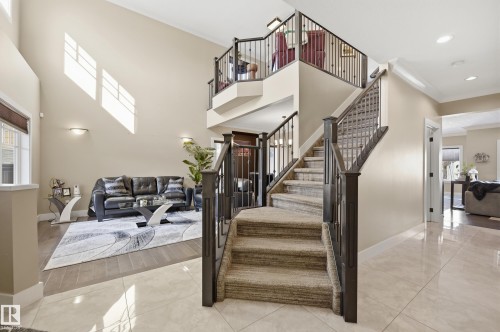 Expansive entryway featuring a grand staircase with carpeted treads and dark wood banisters, complemented by a two-story ceiling and natural light from high windows - 2018 Armitage Green, Edmonton, AB - Indoor Photo Showing Other Room