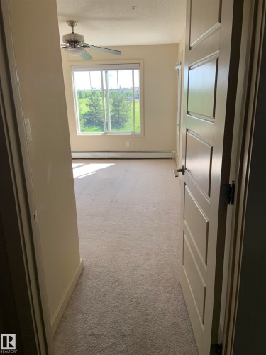 Neutral-toned carpeted interior space featuring a ceiling fan, a large window with white trim, and a three-panel interior door - 307 6084 Stanton Drive, Edmonton, AB - Indoor Photo Showing Other Room