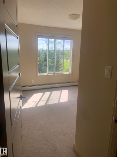 Spacious room featuring a large window, light neutral carpeting, baseboard heating, a contemporary flush-mount ceiling light fixture, and a solid-core paneled door - 307 6084 Stanton Drive, Edmonton, AB - Indoor Photo Showing Other Room