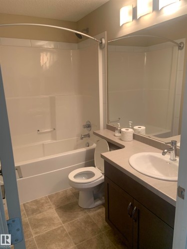 Bathroom featuring a white tub/shower combination with a curved shower rod, a dark wood-finish vanity with a built-in sink, a light-colored countertop, and a wall-mounted light fixture above a mirror - 307 6084 Stanton Drive, Edmonton, AB - Indoor Photo Showing Bathroom