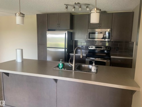 Modern kitchen featuring a breakfast bar with an undermount sink, stainless steel appliances, dark wood-finish cabinetry, a subway tile backsplash, and contemporary pendant lighting - 307 6084 Stanton Drive, Edmonton, AB - Indoor Photo Showing Kitchen With Double Sink With Upgraded Kitchen