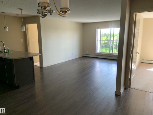 Open-concept living area featuring wood-finish flooring, a large window, contemporary light fixtures, and a kitchen island with a light-toned countertop - 307 6084 Stanton Drive, Edmonton, AB - Indoor