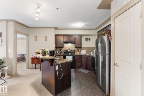The property features a kitchen with dark wood cabinetry, a stainless steel refrigerator, and tiled flooring - #211 4415 48 St, Leduc, AB - Indoor Photo Showing Kitchen