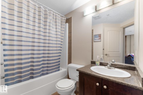 Bathroom featuring a white vanity with a dark countertop and an integrated sink, a toilet, and a shower-tub combination with a striped shower curtain - #211 4415 48 St, Leduc, AB - Indoor Photo Showing Bathroom