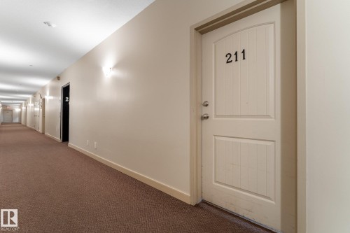 The property features a white paneled door with visible numbering and a metal door handle, set within a neutral-toned hallway with textured brown carpeting - #211 4415 48 St, Leduc, AB - Indoor Photo Showing Other Room