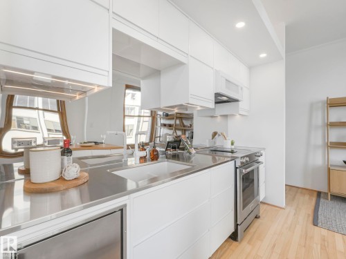 The kitchen features stainless steel countertops, a stainless steel sink with a white faucet, white cabinetry, and stainless steel appliances including an oven, microwave, and dishwasher - 807 10134 100 Street, Edmonton, AB - Indoor Photo Showing Kitchen With Upgraded Kitchen