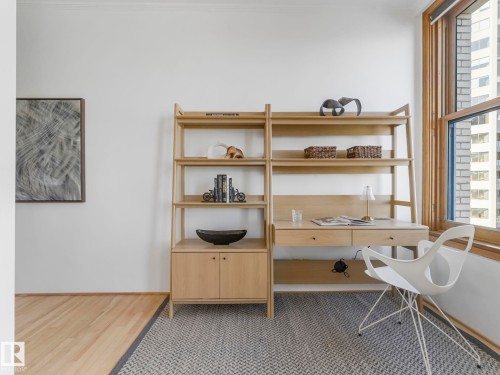 This versatile space features a built-in wooden desk and shelving unit, complemented by wide plank wood flooring and a patterned area rug - 807 10134 100 Street, Edmonton, AB - Indoor