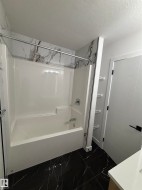 Bathroom featuring a white tub and shower combination with a modern shower head and fixture, dark marble-style tile flooring, and a white interior door with a black handle - 