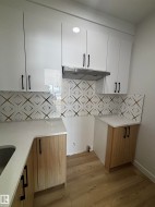 The kitchen features white upper cabinetry with black handles, a patterned backsplash, and light wood-look lower cabinetry - 