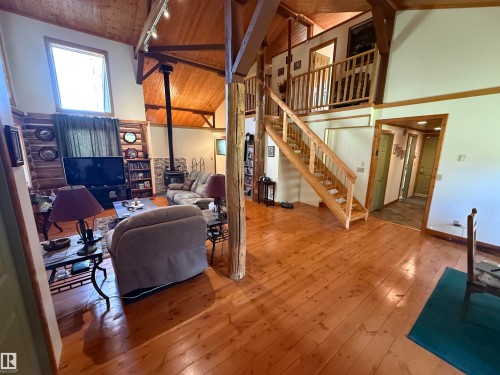 Great room featuring wood-finish flooring, a wood-burning stove with a stacked stone facade, exposed wood beams, and a wood staircase leading to an upper landing - 63218 Range Road 254, Rural Westlock County, AB 