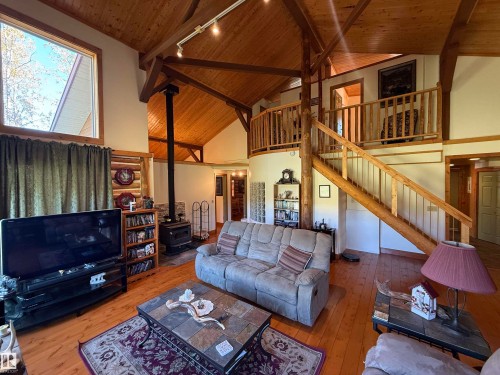 Vaulted wood-paneled ceiling with exposed beams, wood-finish flooring, a freestanding wood-burning stove with stone surround, a prominent wooden staircase, and a second-story loft with a wooden railing - 63218 Range Road 254, Rural Westlock County, AB 